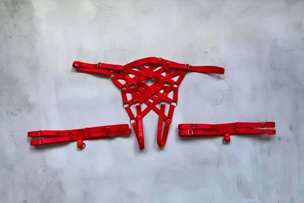 Red caged thong harness with detachable leg bands flat lay showing geometric elastic cage panel and silver O-ring hardware