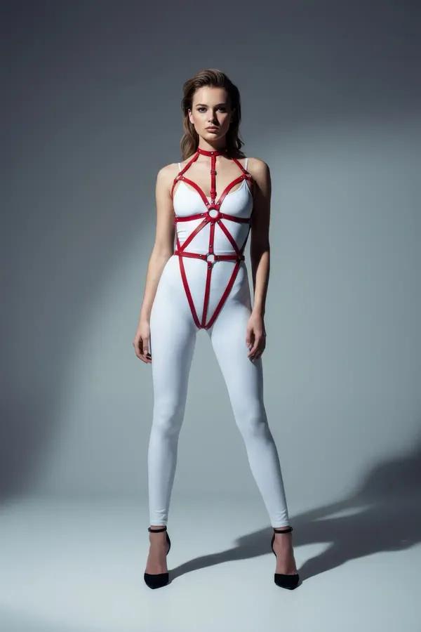 Red faux leather full body harness with radiating geometric straps and silver O-ring detail worn over white bodysuit